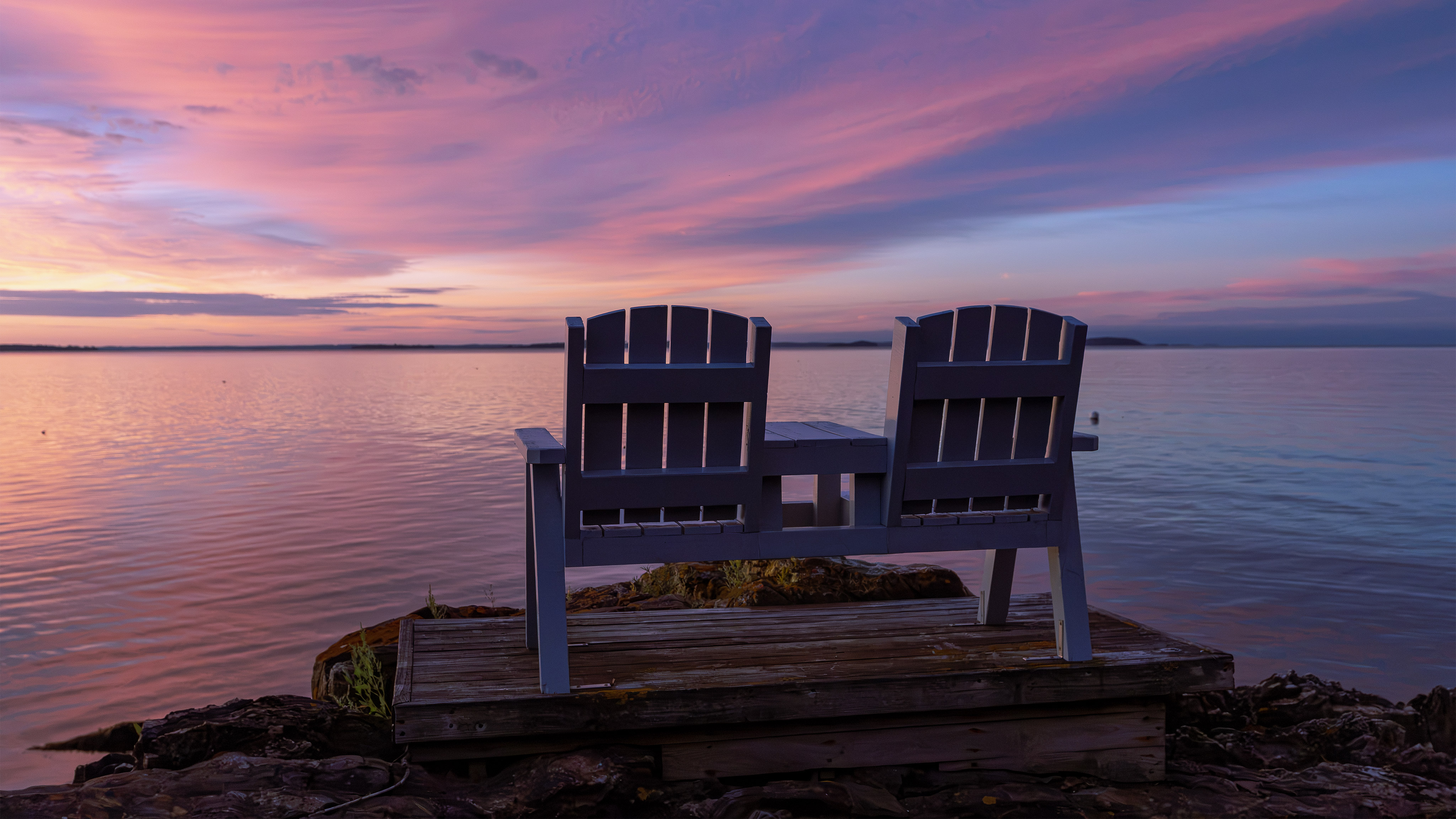 Two Adirondack chairs overlooking the water beneath a pink and purple sunset